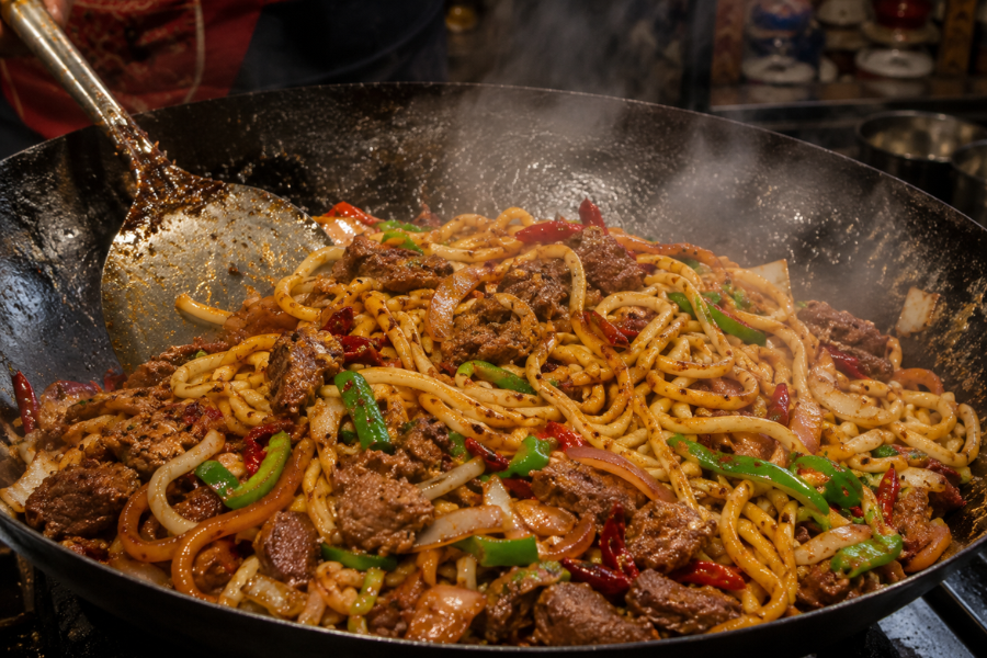 Xinjiang chef cooking dry-fried hand-pulled noodles over high heat in large steel wok, visible flame, noodles slightly charred at edges, strong wok aroma atmosphere, local Xinjiang noodle restaurant, realistic travel food photography, natural kitchen lighting, horizontal image Xinjiang chef cooking dry-fried hand-pulled noodles over high heat in large steel wok, visible flame, noodles slightly charred at edges, strong wok aroma atmosphere, local Xinjiang noodle restaurant, realistic travel food photography, natural kitchen lighting, horizontal image